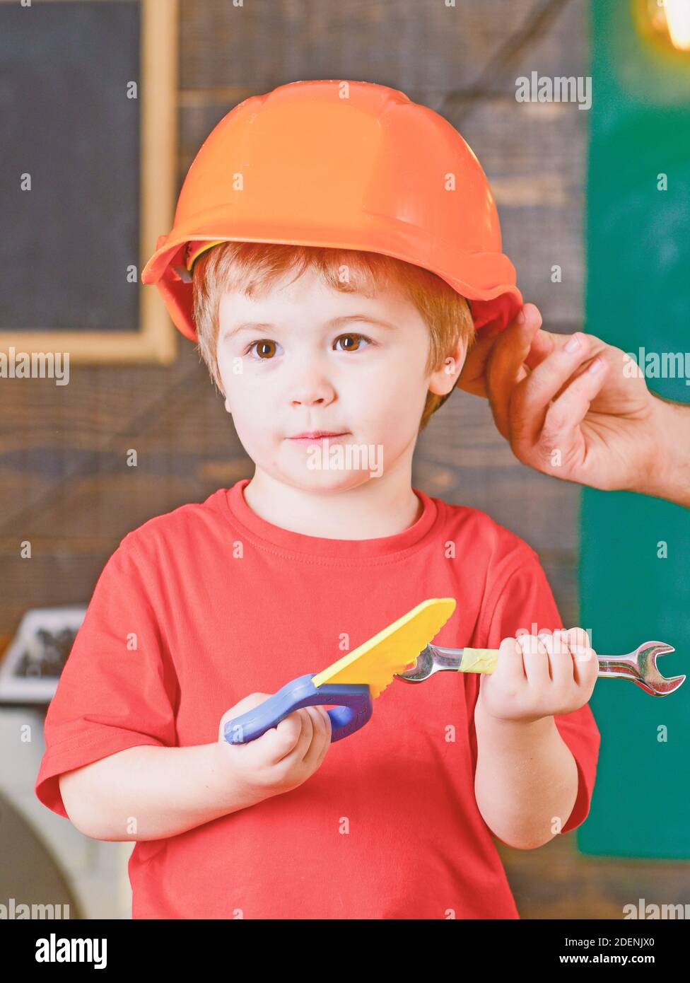 Educational game concept. Kid playing with toy tools. Male hand holding orange protective helmet. Stock Photo