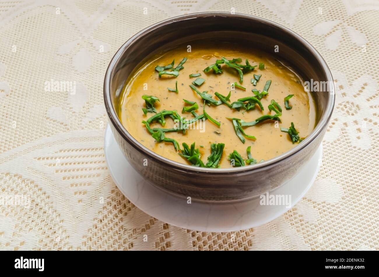 On a lacey background is a brown striped bowl of rich thick meatless pumpkin soup, garnished with green spinach. Stock Photo