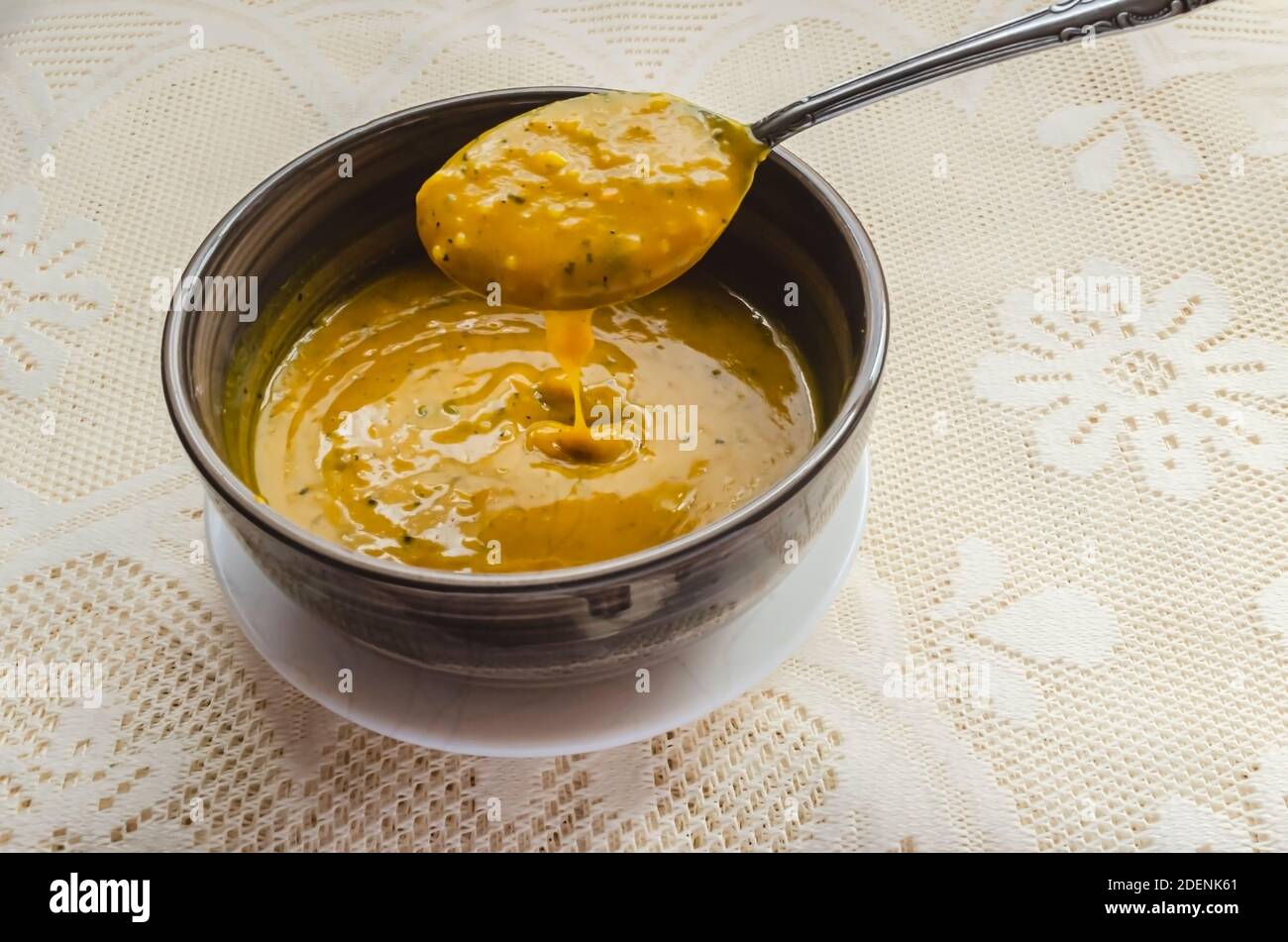 A spoon is lifting thick smooth pumpkin soup from a bowl that is in a small white plate on a lace tablecloth. Stock Photo