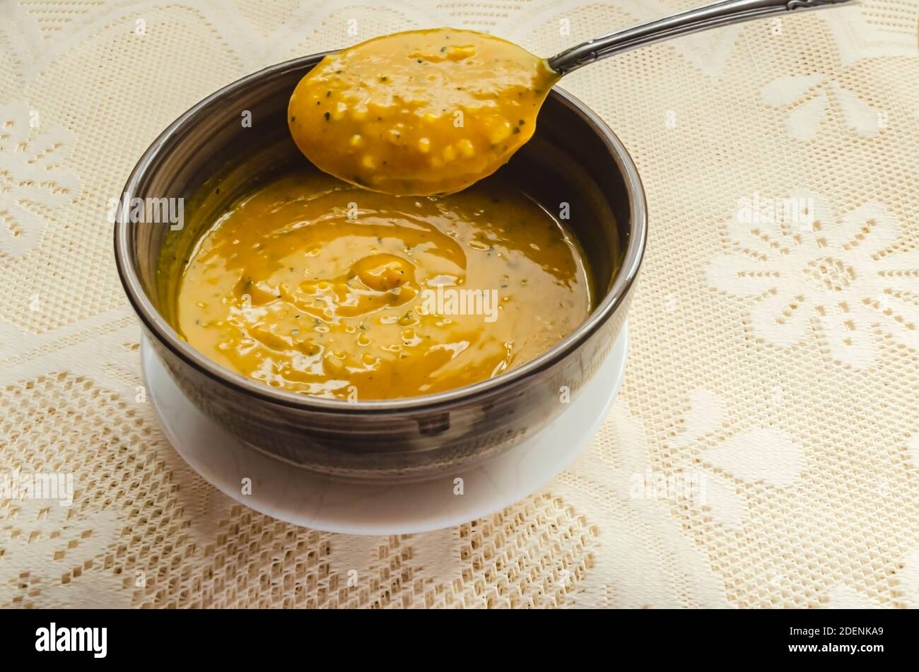 A spoon is lifting thick smooth pumpkin soup from a bowl that is in a small white plate on a lace tablecloth. Stock Photo