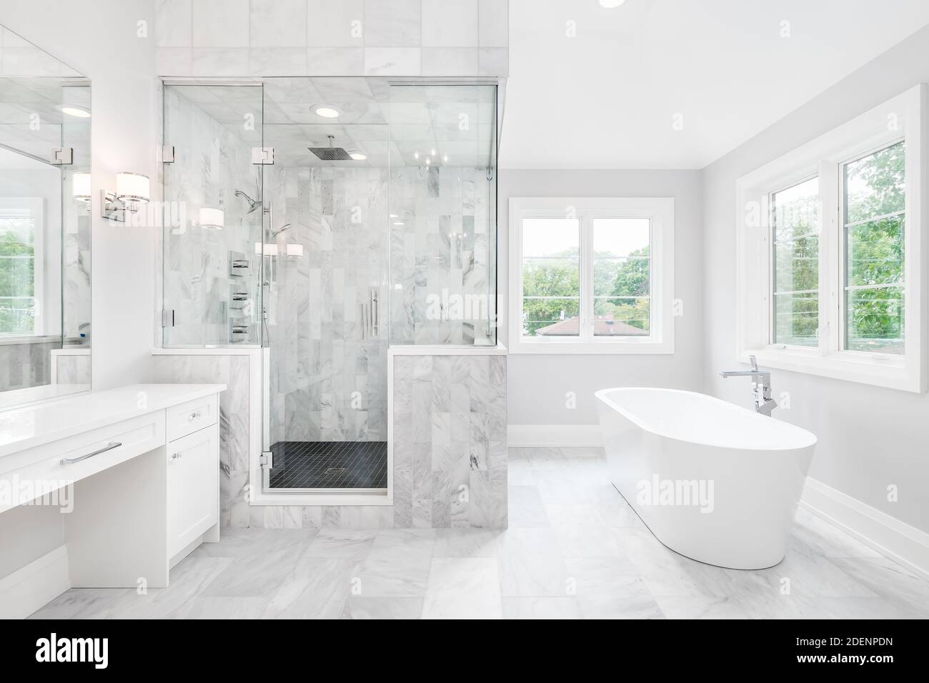 A large luxurious bathroom with a stand alone tub, white vanity, and a glass stand up shower with marble tiles and bench seat. Stock Photo