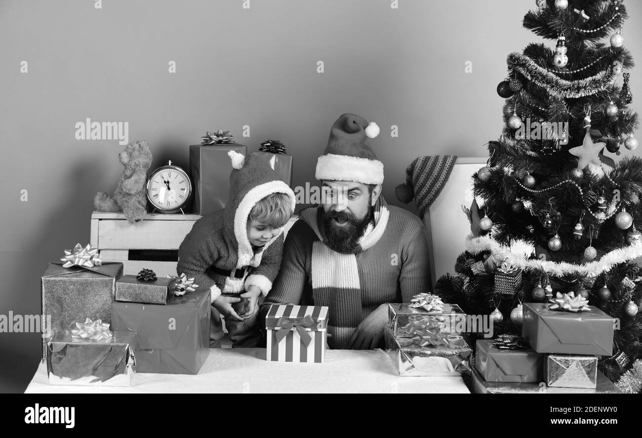 Man with beard and son with shocked faces look at box. Santa and little assistant among gift boxes near Christmas tree. Christmas family opens present Stock Photo
