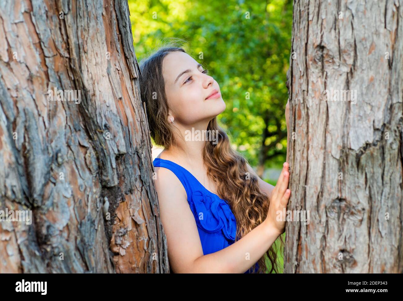 Little kid dreamer with long wavy hair and beauty look dream at tree trunk in summer park, future. Stock Photo