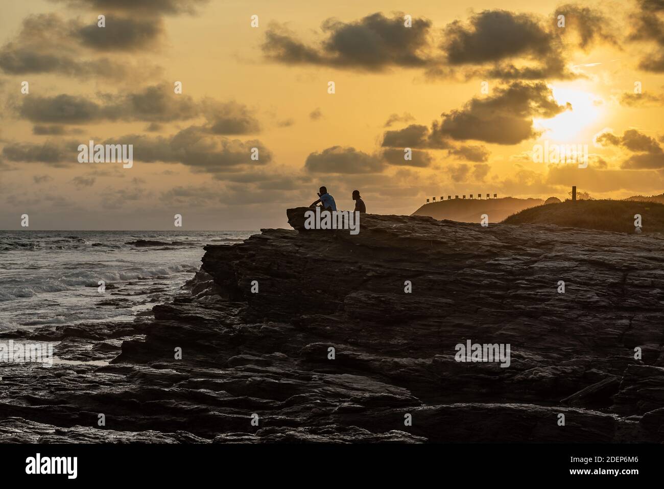 Sunset over Accra beach with rocks and two people sitting and enjoying the sea with hilly ground where you see the shooting range signs in the backgro Stock Photo