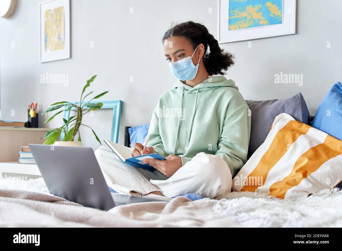 Mixed race teenage girl wearing face mask remote studying online at home. Stock Photo