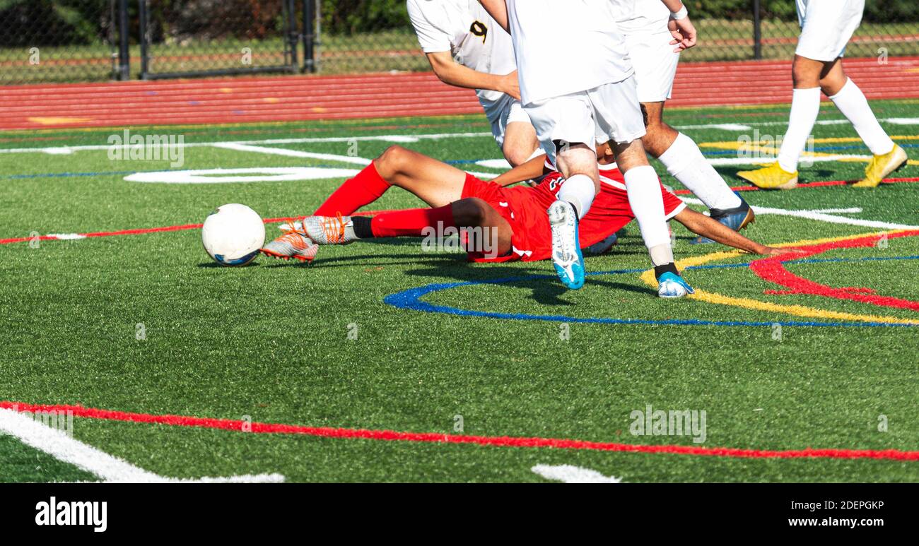 A high school boy soccer player in a red uniform slide tackles the ball away from the competition in white uniforms. Stock Photo