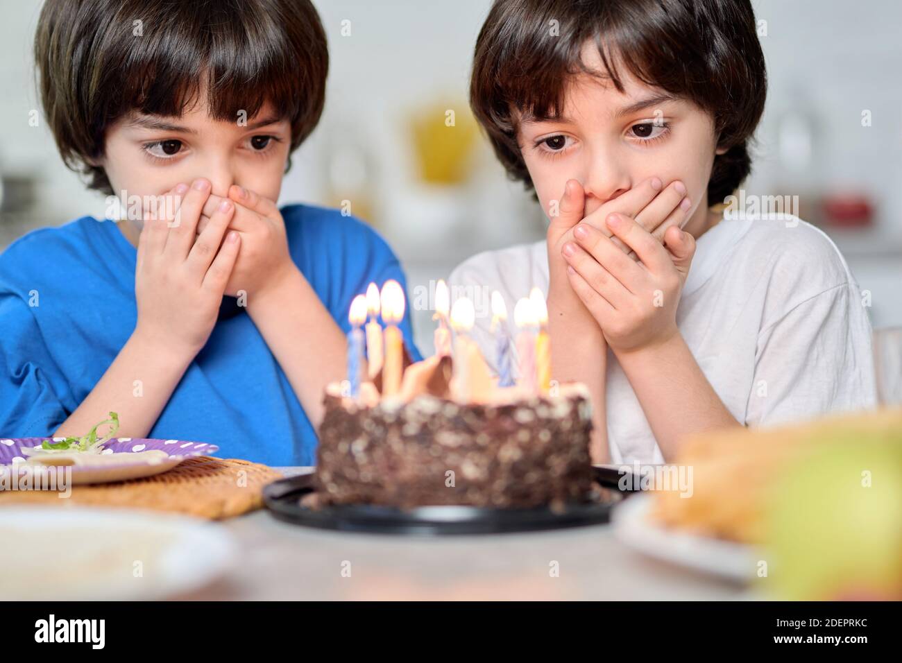 Adorable little hispanic boys looking at birthday cake, making wishes while getting ready for blowing candles, celebrating birthday at home. Selective focus on children Stock Photo