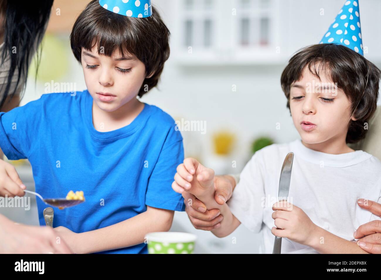 Two cute little brothers in birtday caps eating, having dinner while celebrating birthday together at home. Kids, celebration concept Stock Photo