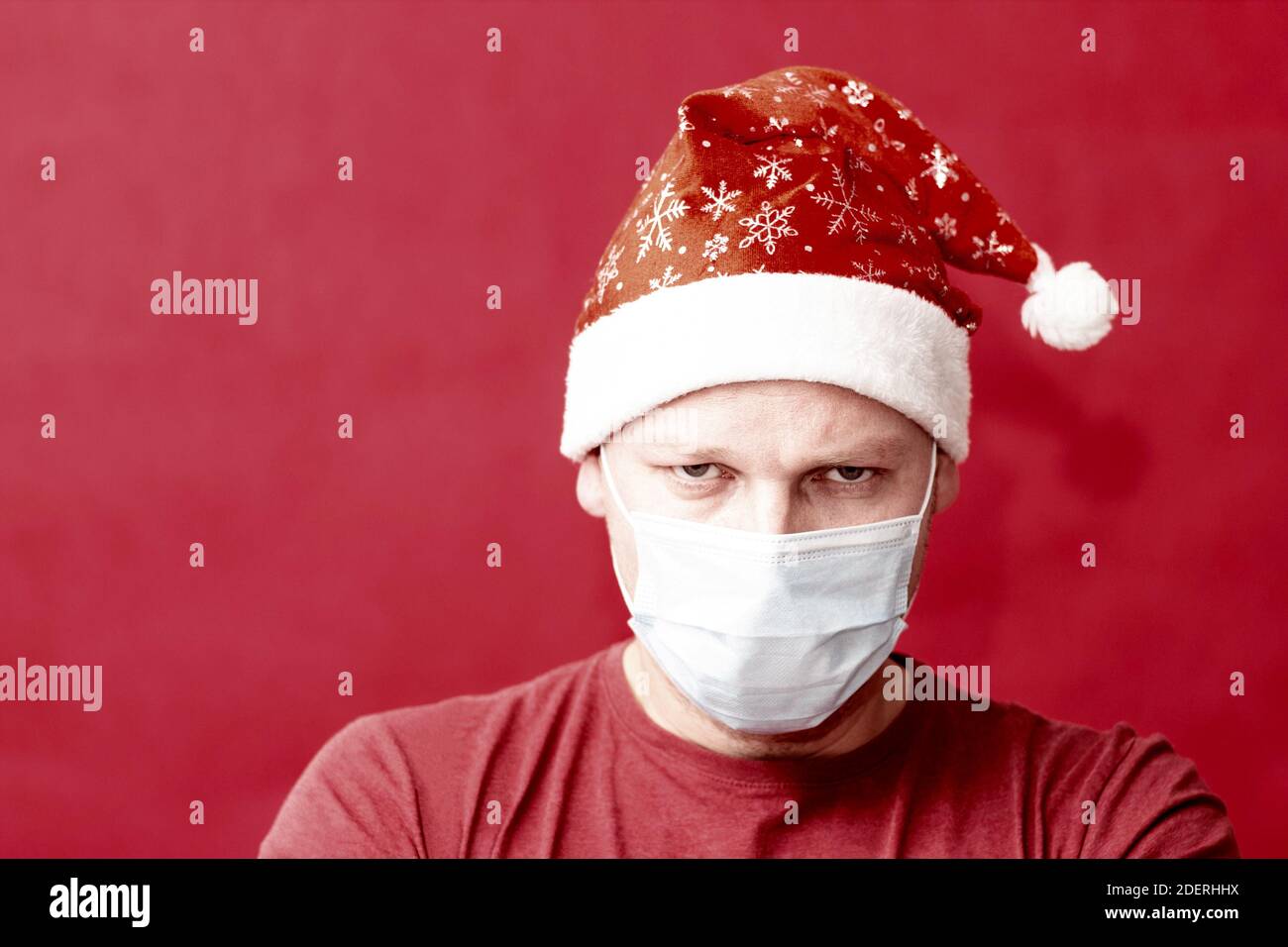 Grumpy man in Santa hat and medical protective mask on a red background. Stock Photo