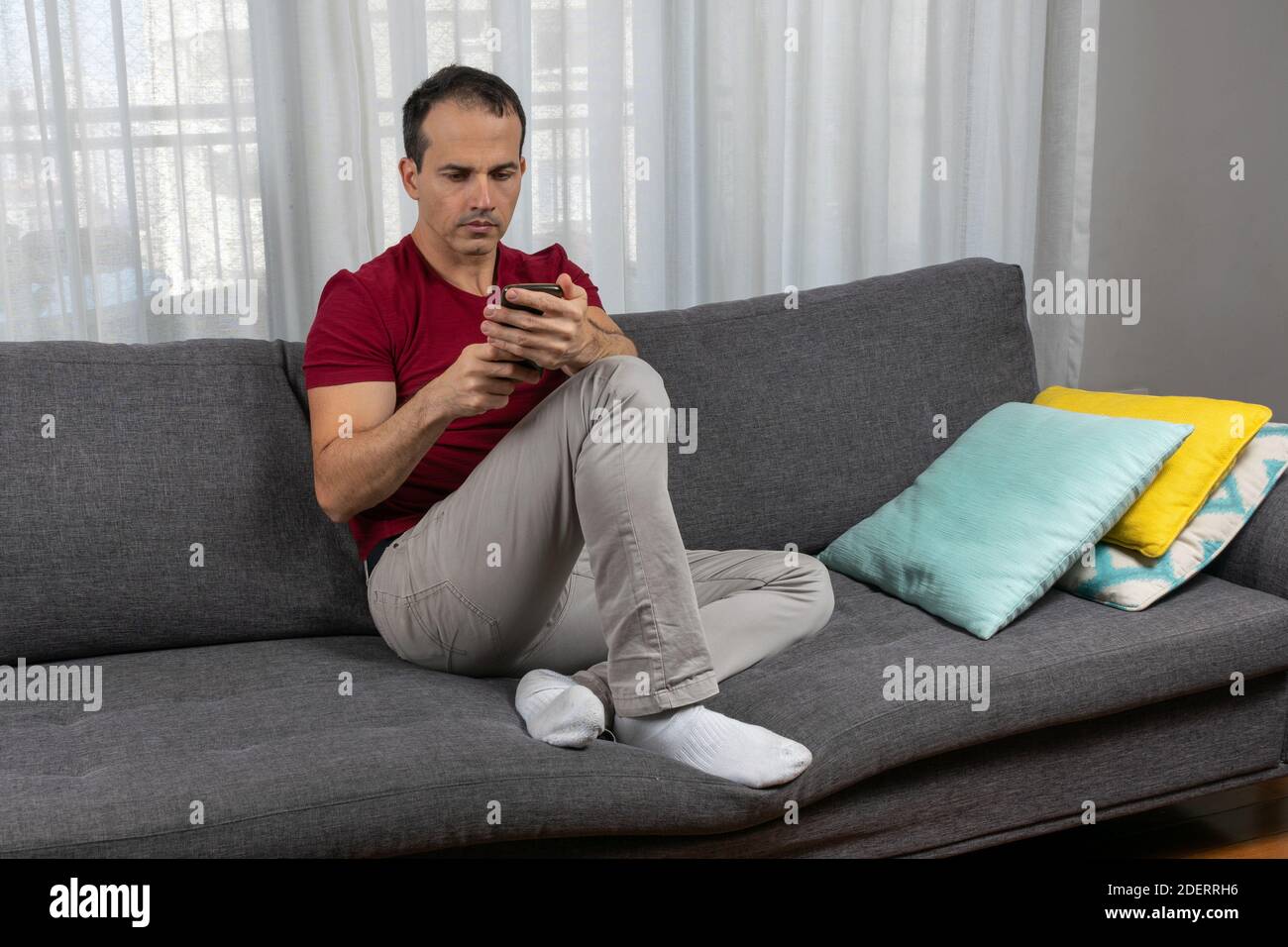 Mature man (44 years old) sitting on the couch and looking mindfully and holding his smartphone. Stock Photo