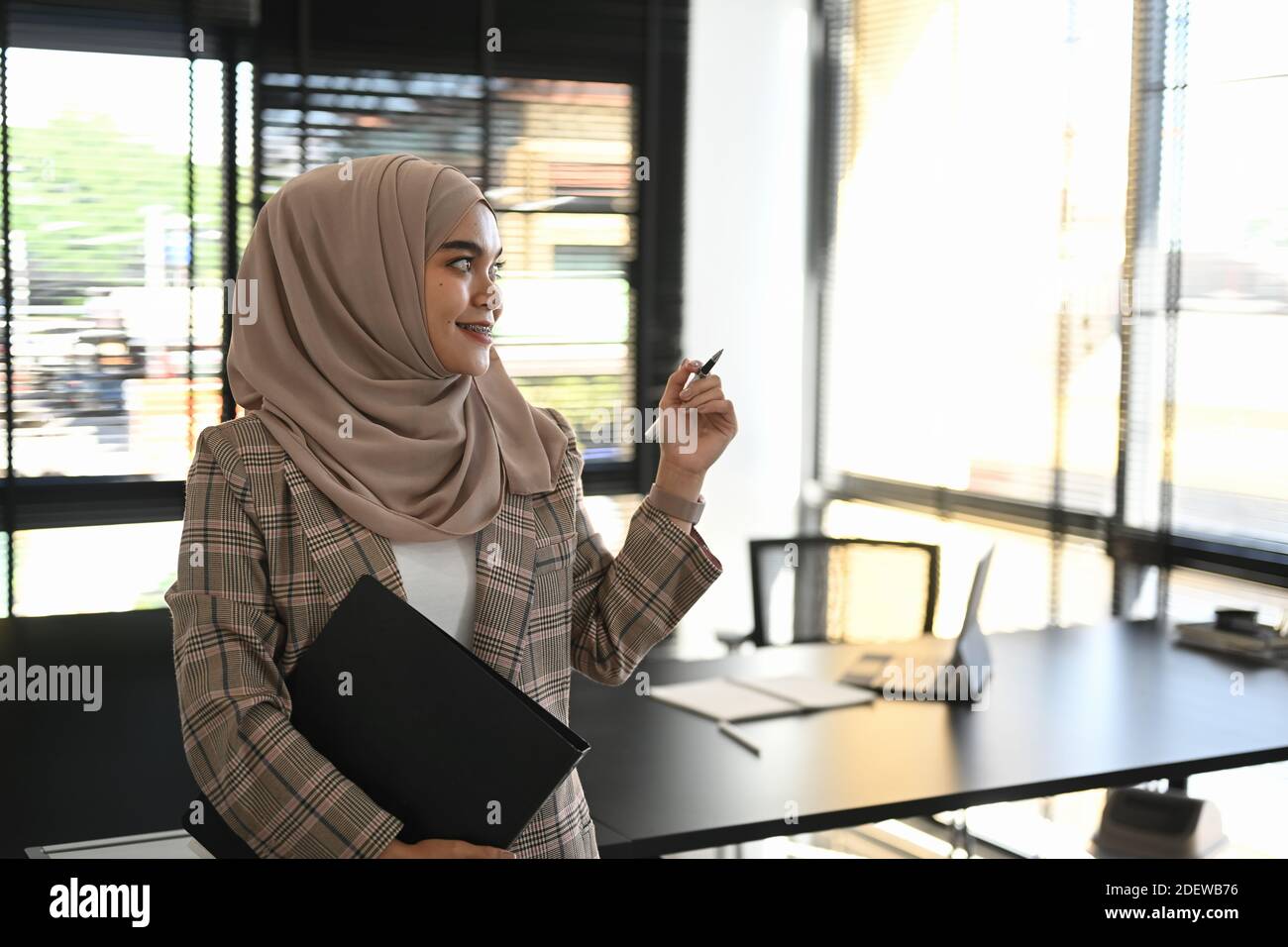 A cheerful muslim woman in abaya standing in office hand holding ...