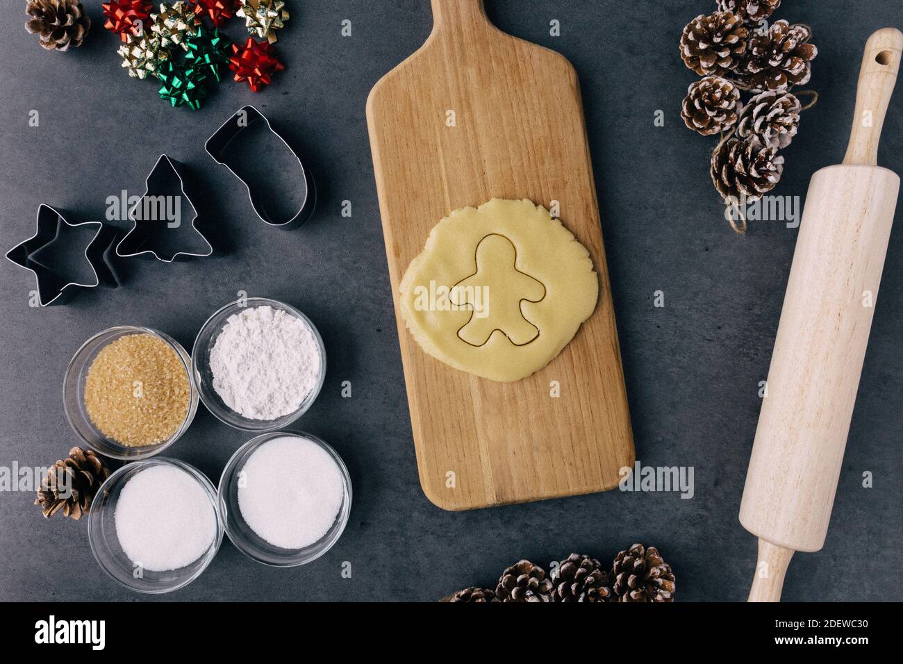 Cutting Christmas Ginger Bread Man Cookie In Raw Cookie Dough With Pine Cones, Holiday Ribbons, And Ingredients on Dark Slate Texture Stock Photo