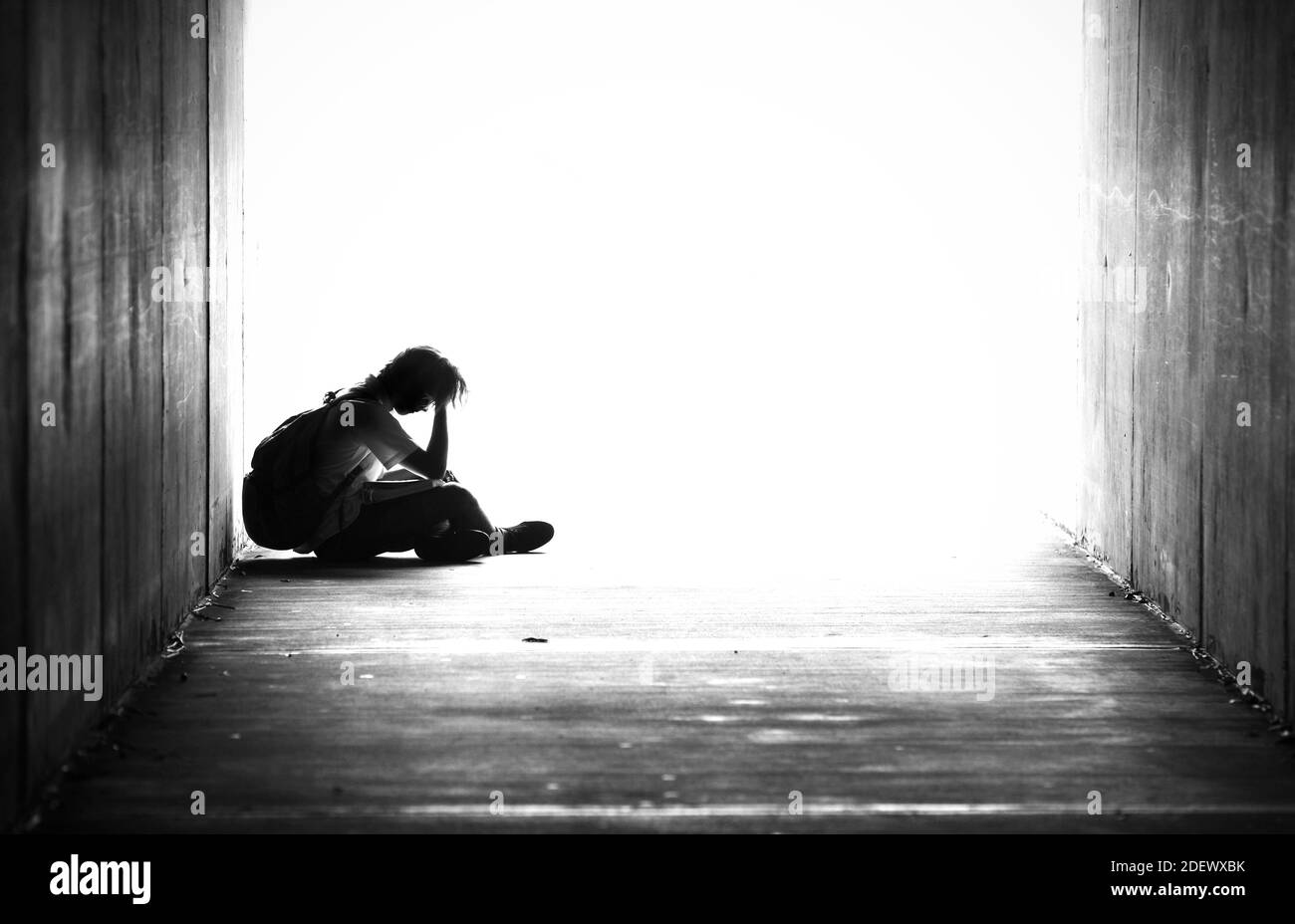 Silhouette of a young male sitting alone in a tunnel with his head in his hands. Black and white image of school kid with bag on his bag. Down and out Stock Photo