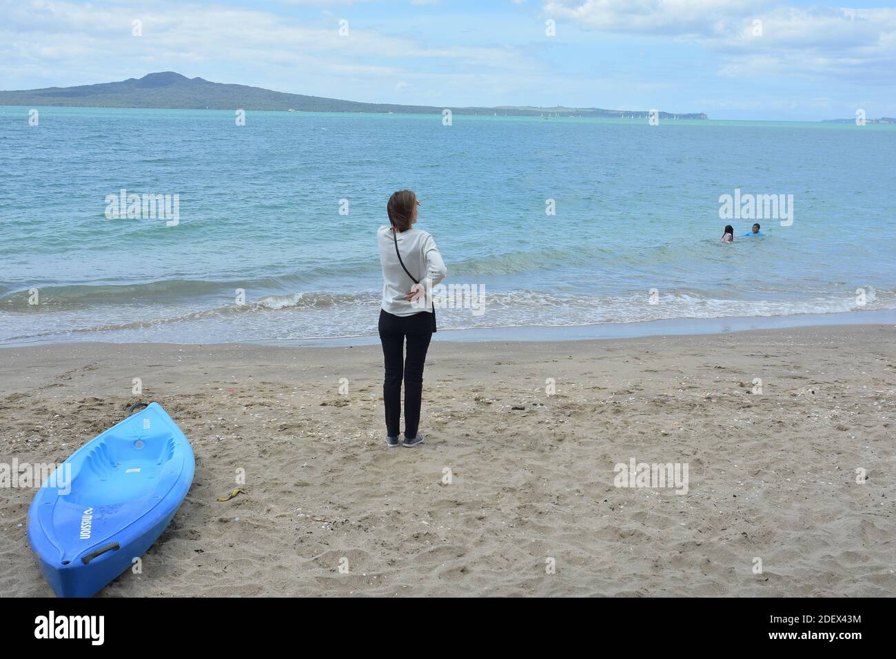 AUCKLAND, NEW ZEALAND - Nov 21, 2020: View of woman standing and watching swimming kids at Mission Bay beach with Rangitoto Island in background and b Stock Photo