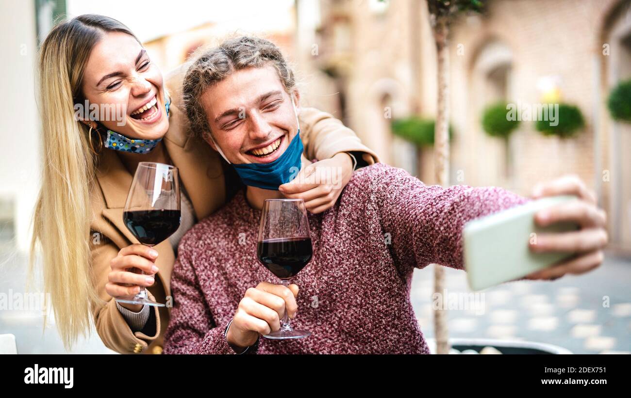 Young couple in love with open face mask taking selfie at outside wine bar - Happy millennial lovers enjoying lunch together at restaurant patio Stock Photo