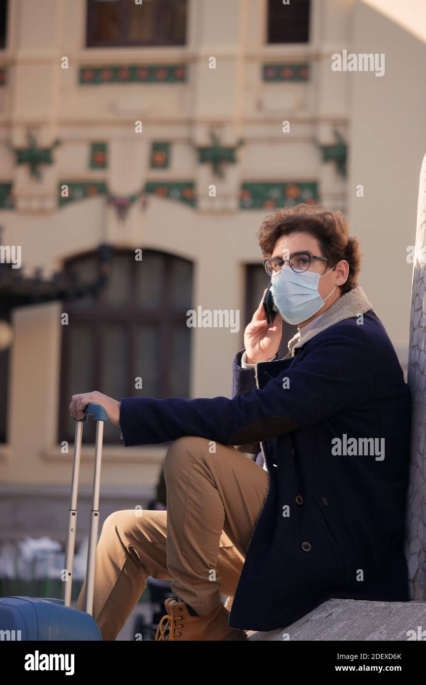 Close up recognizable young man with a mask on sitting outside the train station with his suitcase calling a taxicab. Business trip 2020. Stock Photo