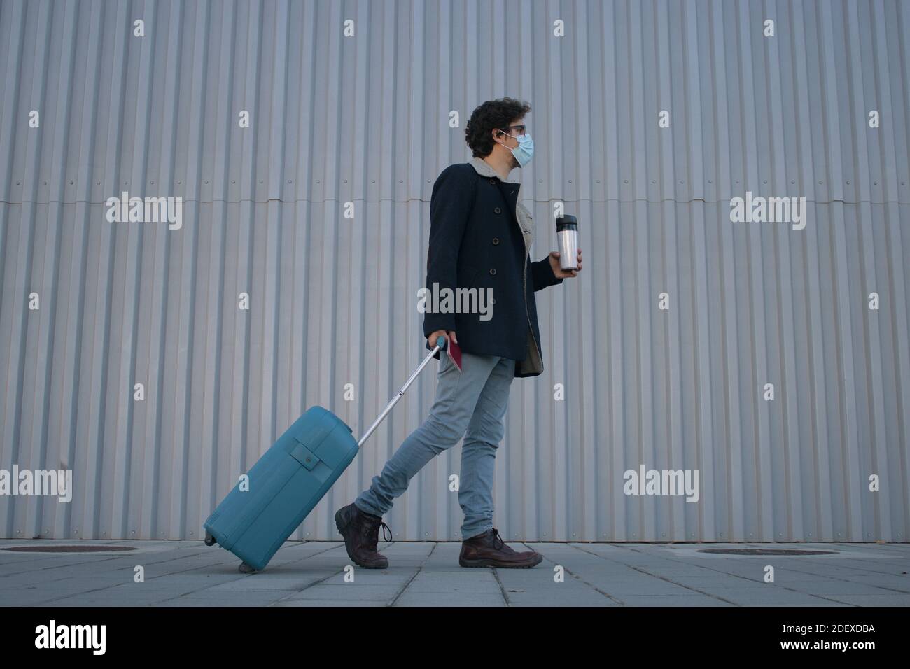 Man with suitcase and passport in his hand. He walks straight to the airport with a take away coffee during Covid 19. Stock Photo