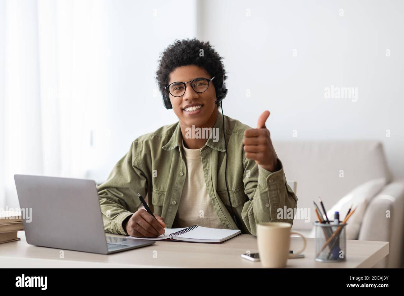 Positive black student in headset gesturing thumb up near laptop at home Stock Photo
