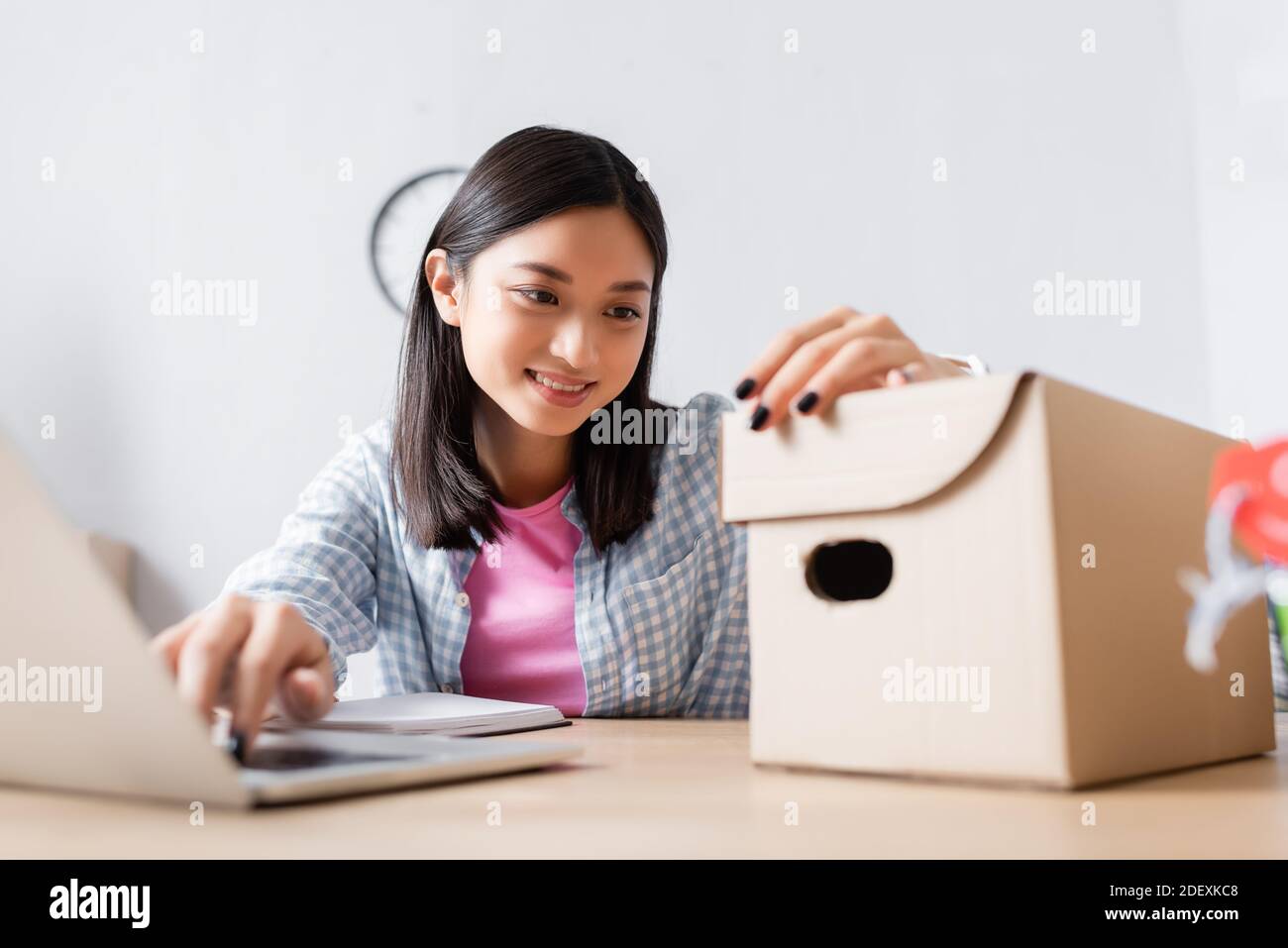 Happy asian volunteer looking at carton box while sitting at desk in charity center on blurred foreground Stock Photo