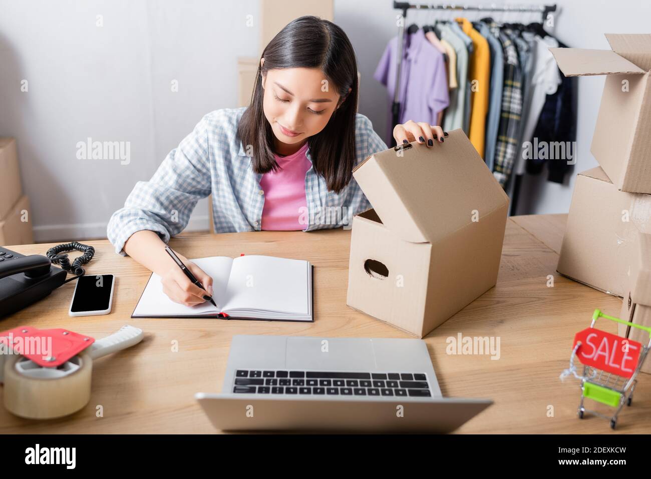 Asian volunteer opening carton box and writing in notebook at desk with digital devices in charity center Stock Photo