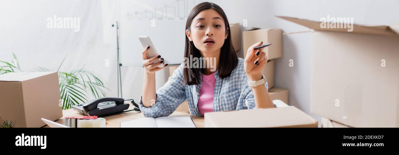 Asian volunteer with smartphone pointing with pen at carton box in charity center, banner Stock Photo