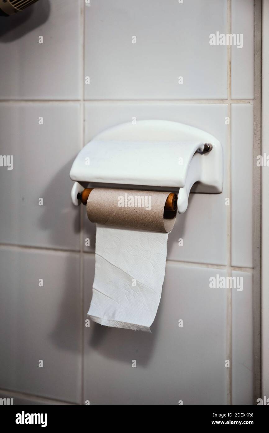 One single sheet of toilet paper is left on an empty toilet paper roll hanging on a white tiled wall in a bathroom. Stock Photo