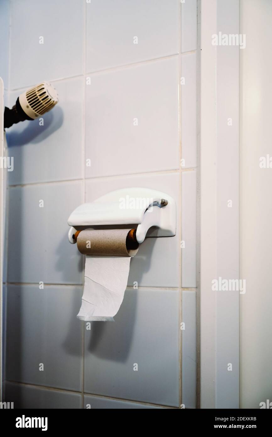 One single sheet of toilet paper is left on an empty toilet paper roll hanging on a white tiled wall in a bathroom. Stock Photo