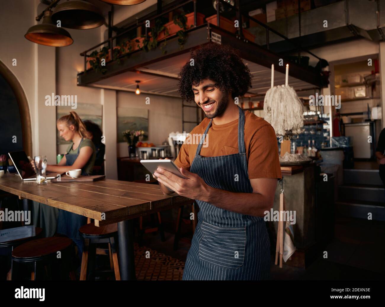 Male waiter with curly hair working in cafe wearing apron with using digital tablet with female working in background on laptop Stock Photo