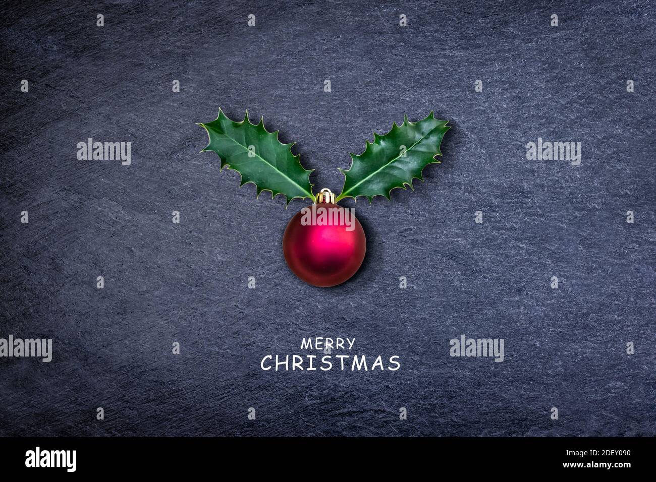 Christmas card - Reindeer face made of Holly leaves and red bauble on dark stone. Stock Photo