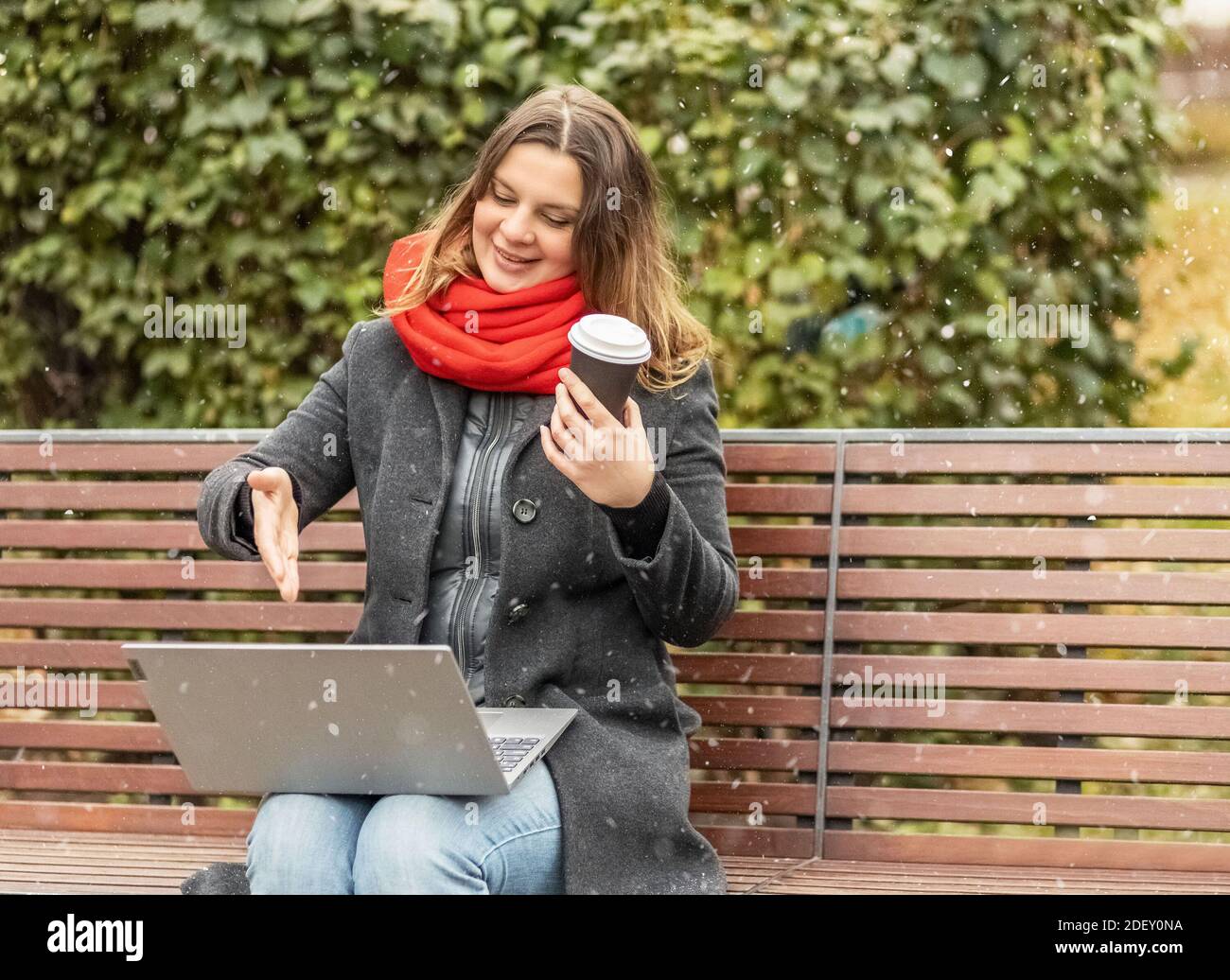 Young woman working on laptop sitting on a park bench with coffee. Computer and online shopping use. Distance learning, online education Stock Photo