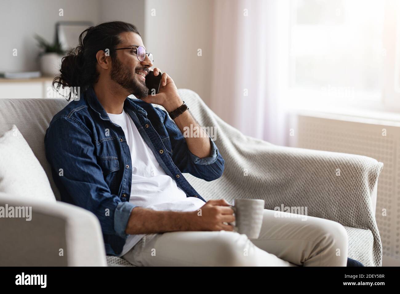 Smiling Indian Guy Resting At Home, Talking On Cellphone And Drinking Coffee Stock Photo