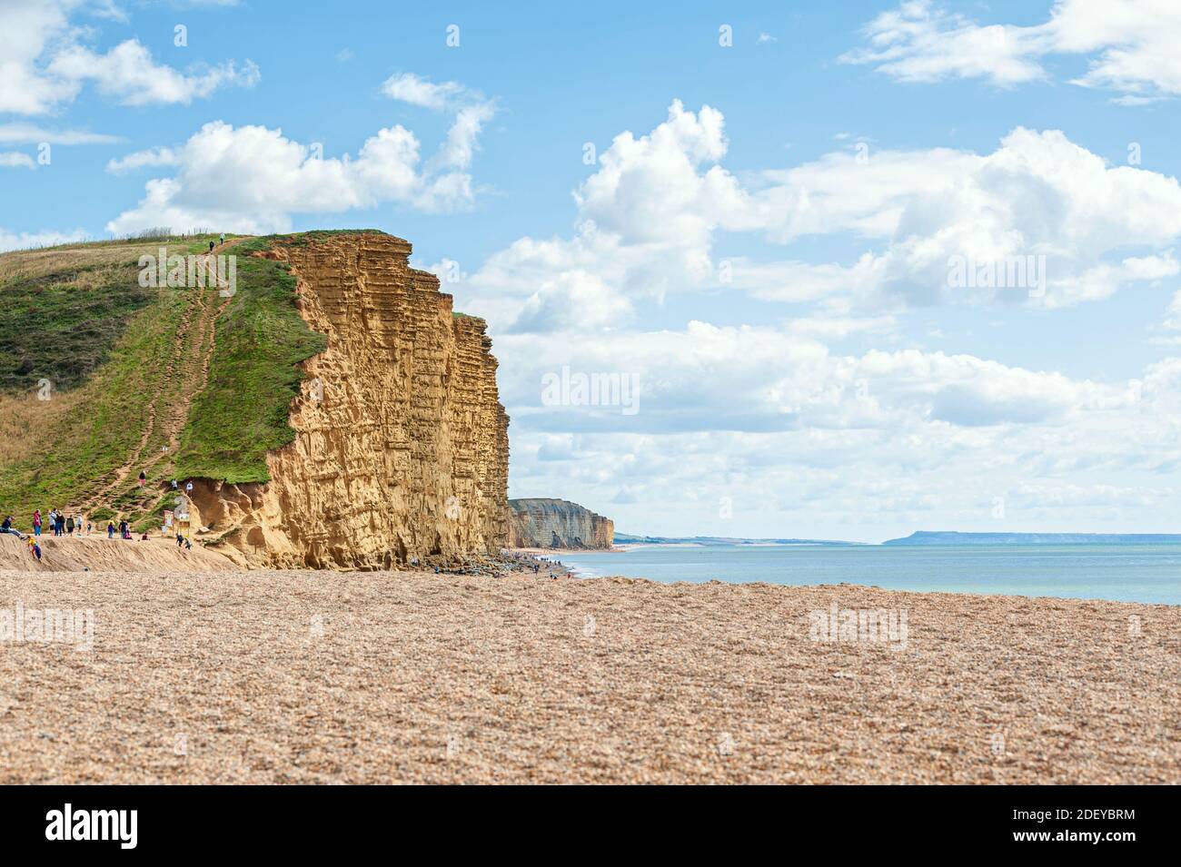 People walking on golden beach underneath towering cliffs on sunny summer day. Jurrasic coastline of West Bay in Dorset. UK Stock Photo