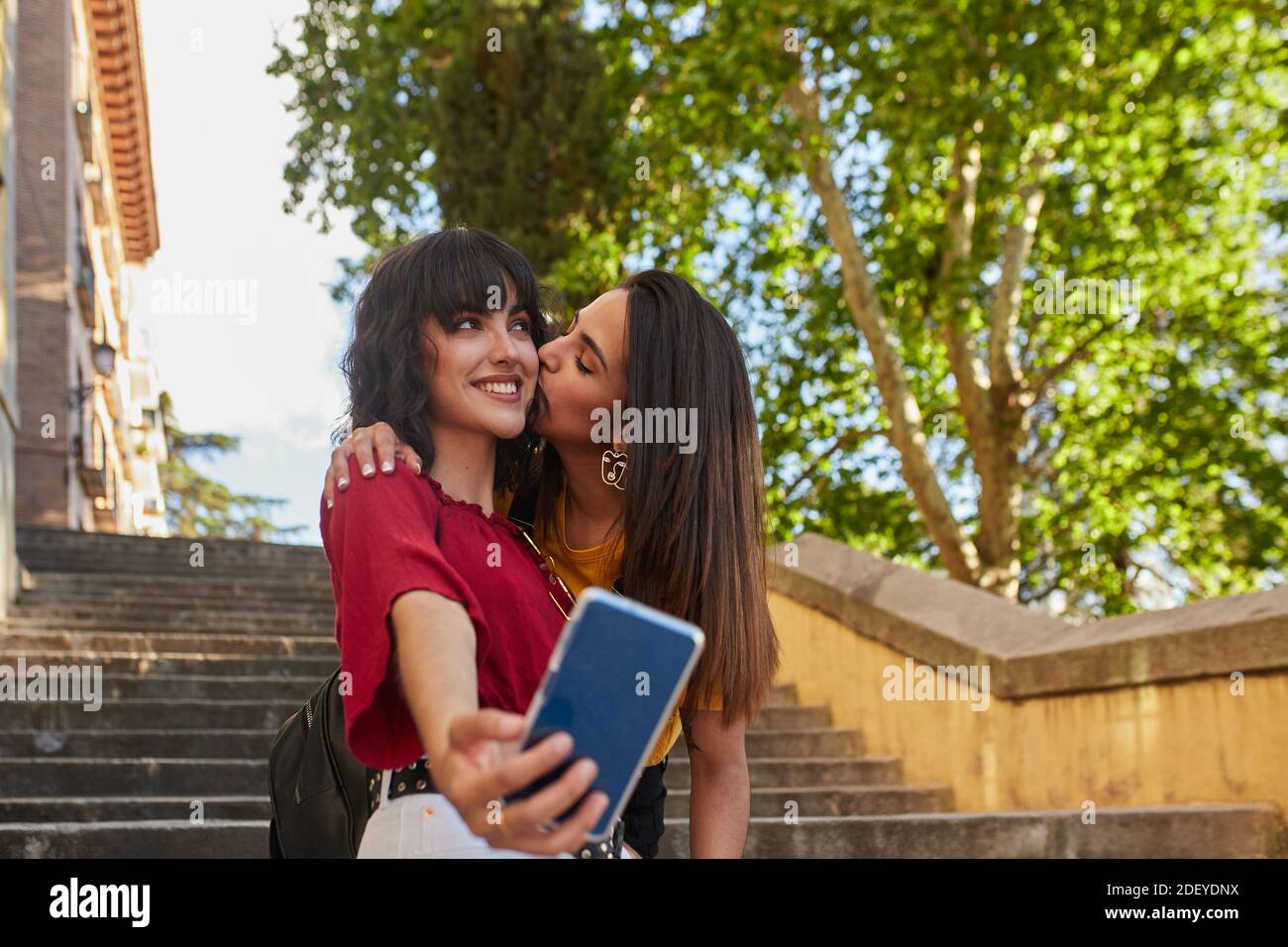 Stock photo of two teenager girls standing stairs and taking a selfie. One of them is kissing the other They are wearing casual cloth and sunglasses. Stock Photo