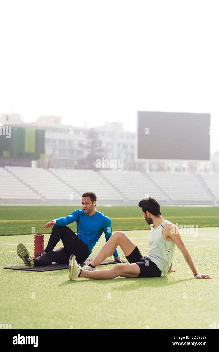 Stock photo of caucasian and black male athletes sitting on the track while stretching their legs and arms. Stock Photo
