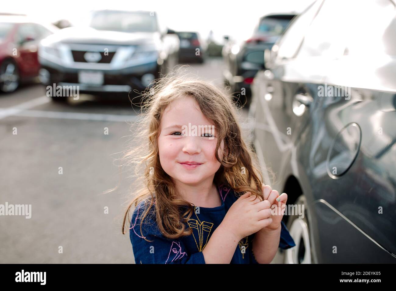 Smiling 3 yr old girl twining long hair standing in parking lot Stock Photo