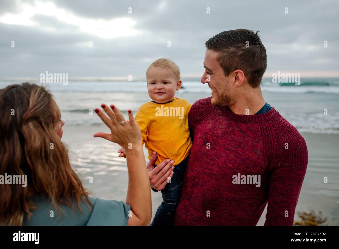 Mom high-fives baby held by dad in red sweater at the ocean Stock Photo