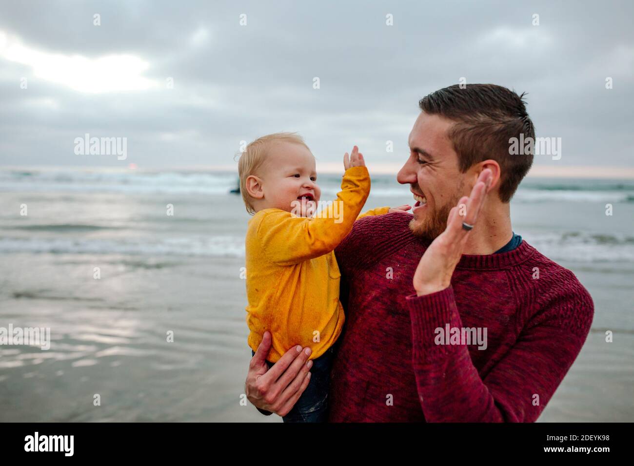 Laughing 30 yr old father holding baby at the ocean giving high-fives Stock Photo