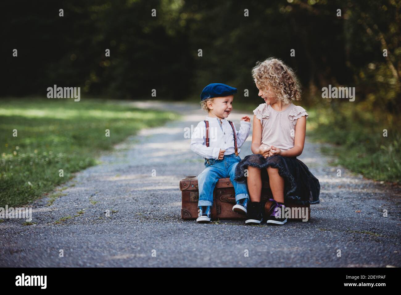 Brother and sister sitting on a vintage suitcase smiling at each other Stock Photo