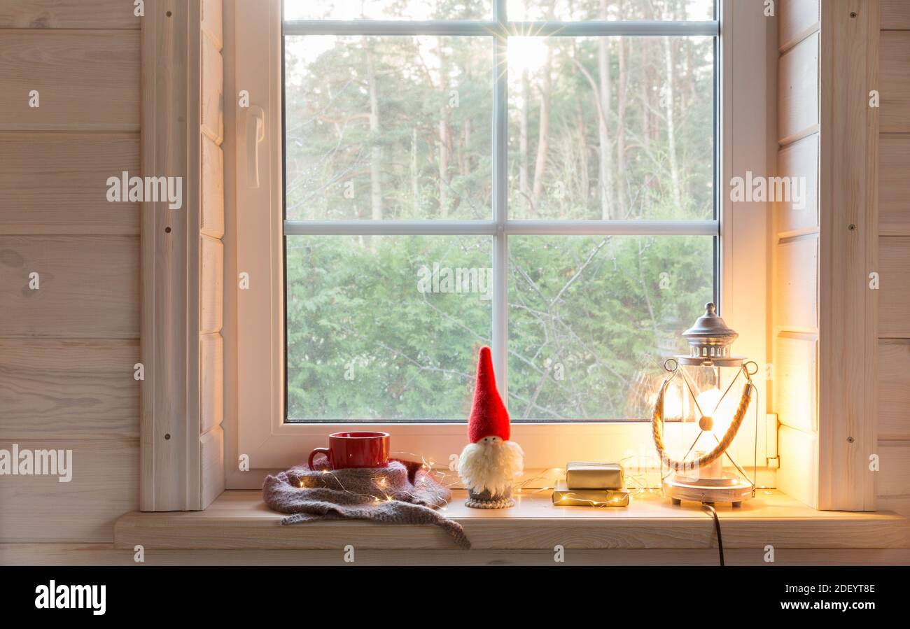 Christmas lantern, Christmas gnome,and red mug on the window of a wooden house overlooking the winter garden. Stock Photo
