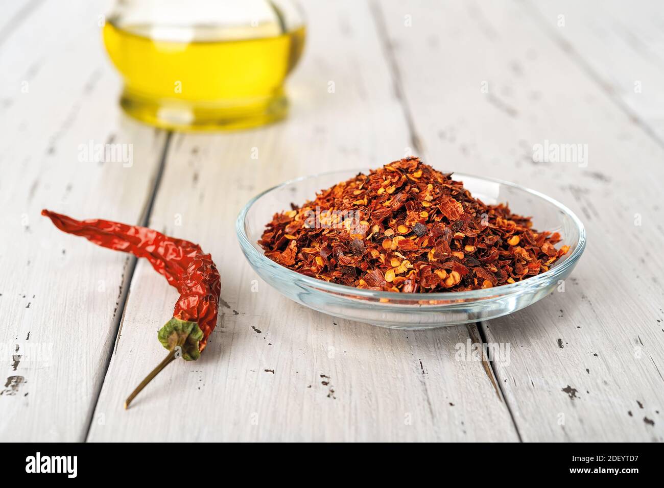 Red hot pepper flakes in a glass saucer and dry wrinkled pepper pod on a rustic wood table. Natural spices and seasonings for meat, fish and vegetable Stock Photo