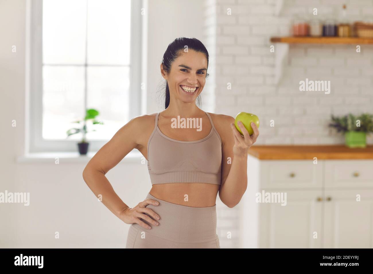 Fit woman standing in the kitchen, holding fresh green apple and smiling at camera Stock Photo