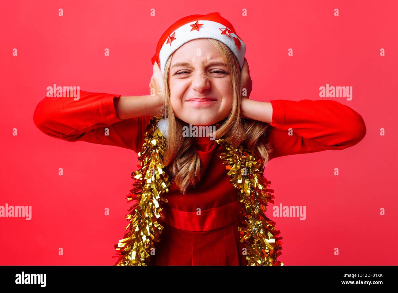 Portrait of a teenager in a Santa hat and tinsel on the neck, screaming and covering the ears of the girl, wants to hear nothing . Christmas, human em Stock Photo