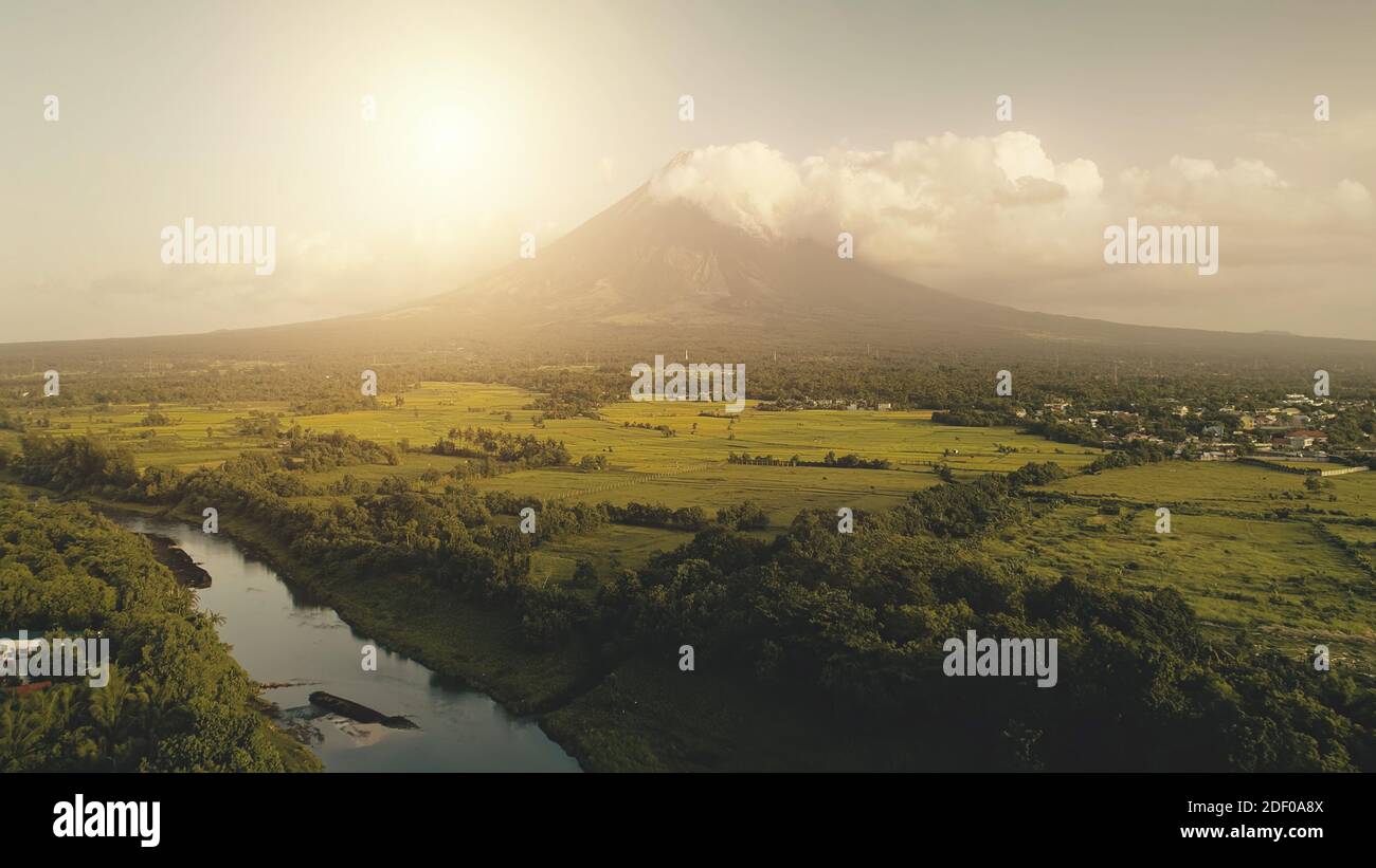 Tropic sun shine over river at erupt volcano aerial. Fields, meadows at nobody nature landscape. Green grass valley at banks of tropical stream at Philippines countryside. Cinematic Mayon landmark Stock Photo