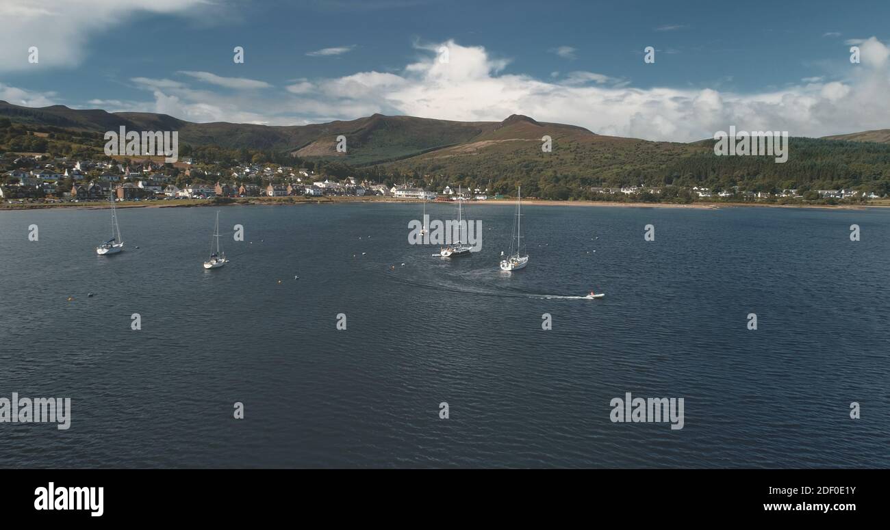 Yacht racing at ocean bay coast aerial. Passengers on sailboat at open sea at summer cloudy day. Cinematic scenery of luxury cruise on sail boat. Brodick harbor at mountain island of Arran, Scotland Stock Photo