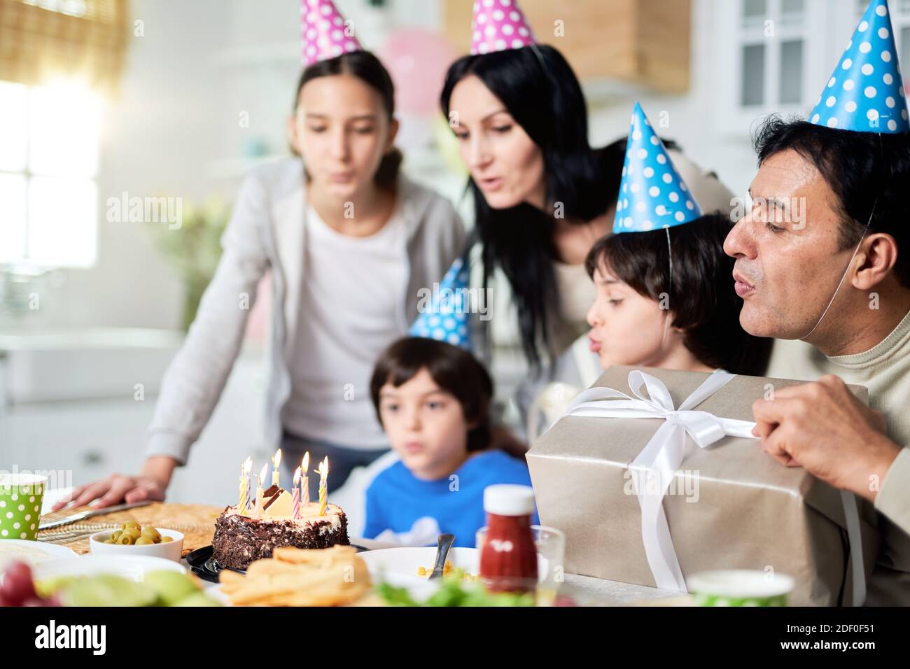 Close latin family with children wearing birthday caps, blowing candles on a cake while celebrating birthday together at home. Selective focus Stock Photo