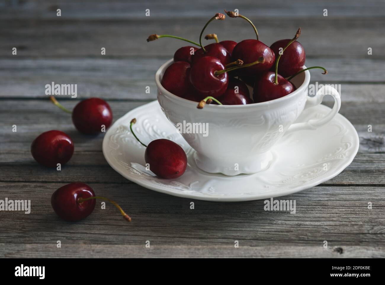 red cherries in white tea cup on aged wooden background, fruit still life Stock Photo