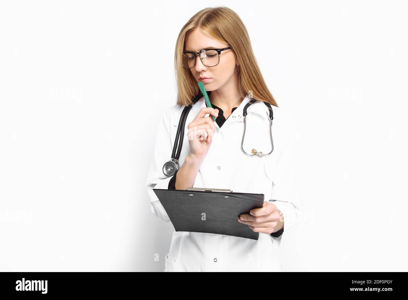 A pensive doctor, a female intern with glasses, with a stethoscope around his neck, writes the diagnosis in his folder on a white background. medicine Stock Photo
