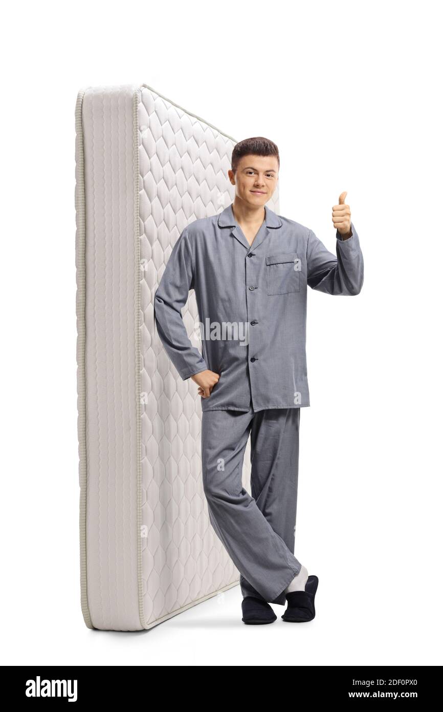 Full length portrait of a guy in pajamas leaning on a mattress and showing thumbs up isolated on white background Stock Photo