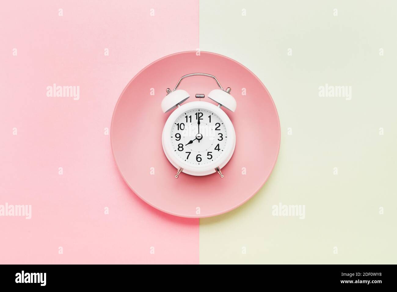 Intermittent fasting concept. White alarm clock on the empty pink plate on the pink-green background. Top view, copy space for text Stock Photo