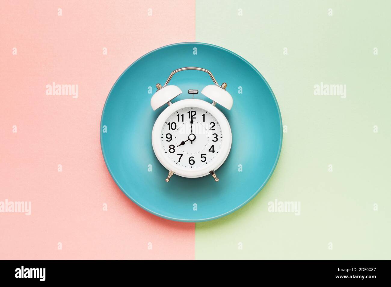 Intermittent fasting concept. White alarm clock on the empty blue plate on pink-green. Eight-hour feeding window concept. Flat lay, copy space. Stock Photo